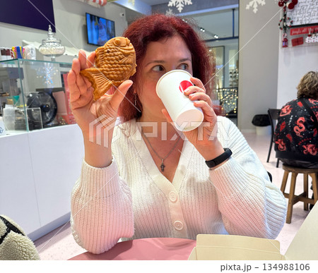 Woman smiling while holding fish shaped taiyaki pastry and warm drink. Playful winter snack, holiday cafe mood, joyful indulgence, cozy atmosphere, and cheerful seasonal lifestyle moment indoors. 134988106