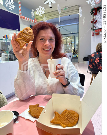 Woman smiling while holding fish shaped taiyaki pastry and warm drink. Playful winter snack, holiday cafe mood, joyful indulgence, cozy atmosphere, and cheerful seasonal lifestyle moment indoors. 134988155