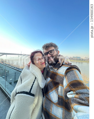 Couple taking selfie together near river embankment in city. Emotional closeness, shared happiness, partnership warmth, everyday romance, and authentic lifestyle moment outdoors. Couple taking selfie together near river embankment in city. Emotional closeness, shared happiness, partnership warmth, everyday romance, and authentic lifestyle moment outdoors. 134988163