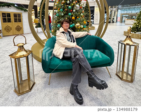 Woman sitting on sofa near decorated Christmas tree installation. Seasonal interior design, festive public space, winter comfort, calm leisure, and lifestyle pause within holiday shopping environment. Woman sitting on sofa near decorated Christmas tree installation. Seasonal interior design, festive public space, winter comfort, calm leisure, and lifestyle pause within holiday shopping environment. 134988179