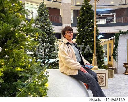 Woman resting near Christmas festive decorated trees in shopping mall interior. Winter holiday atmosphere, public leisure, seasonal decoration, calm enjoyment, and quiet presence during festive urban Woman resting near Christmas festive decorated trees in shopping mall interior. Winter holiday atmosphere, public leisure, seasonal decoration, calm enjoyment, and quiet presence during festive urban 134988180