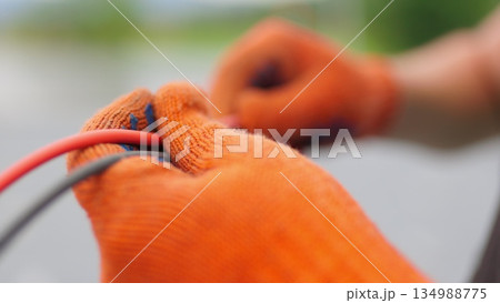 An electrician worker holds a red and black high-voltage electric cable, checks the quality of the cables. Connecting electrical cables. Close up of male hands in protective gloves holding cable. An electrician worker holds a red and black high-voltage electric cable, checks the quality of the cables. Connecting electrical cables. Close up of male hands in protective gloves holding cable. 134988775