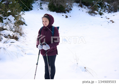 Woman Hiking in Snowy Landscape With Trekking Poles on Winter Adventure Through Peaceful Forest Trail 134989503