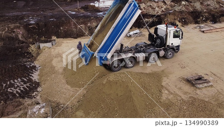 Dump truck unloading soil or sand at construction site. Truck laden with dirt and soil travels in a road construction complex site. 134990389