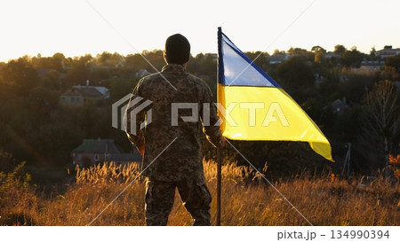 Soldier of ukrainian army going with lifting blue-yellow banner at sunset. Young male military in uniform walks with waving flag of Ukraine at countryside. Victory against russian aggression concept 134990394