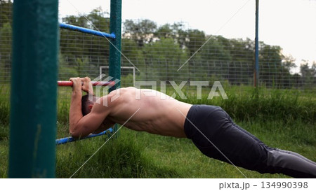Young muscular athlete doing exercise on triceps at sport equipment outdoor. Male shirtless athlete working out at sport ground. Strong sporty man training at nature. Concept of healthy lifestyle Young muscular athlete doing exercise on triceps at sport equipment outdoor. Male shirtless athlete working out at sport ground. Strong sporty man training at nature. Concept of healthy lifestyle 134990398