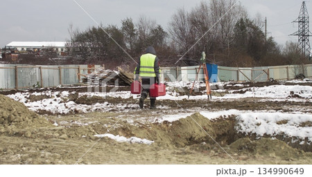 Worker Professional Surveying Engineer At the construction site in his hands two red suitcases. Worker using a theodolite geodetic optical device to measure angles in the horizontal and vertical 134990649