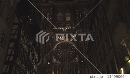 Strasbourg, France - May 8, 2020: Forward push-in camera tilt up from entrance to tower of lighted Notre Dame Cathedral front facade in Petite France old town at night between buildings. 134990664