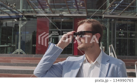 Good-looking confident purposeful adult bearded businessman in glasses in stylish suit going down on steps while leaving the office building and looking at camera. Good-looking confident purposeful adult bearded businessman in glasses in stylish suit going down on steps while leaving the office building and looking at camera. 134990765