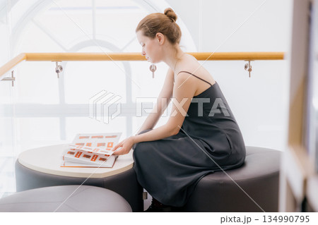 woman in library is reading book sitting on wide chair. lady in black dress flips through pages. woman in library is reading book sitting on wide chair. lady in black dress flips through pages. 134990795