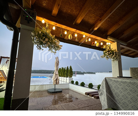 Outdoor terrace with hanging lights and flower decor during rainy evening. Cozy hospitality, calm atmosphere, slow living, seasonal relaxation, and intimate dining space under wooden roof Outdoor terrace with hanging lights and flower decor during rainy evening. Cozy hospitality, calm atmosphere, slow living, seasonal relaxation, and intimate dining space under wooden roof 134990816