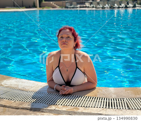 Woman leaning on swimming pool edge under sunlight. Summer relaxation, resort leisure, water enjoyment and calm vacation moment Woman leaning on swimming pool edge under sunlight. Summer relaxation, resort leisure, water enjoyment and calm vacation moment 134990830