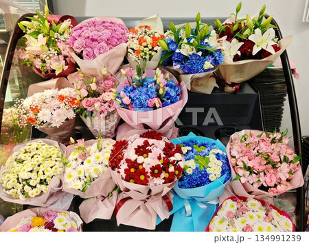 Flower Bouquets Shop Display. Multiple wrapped flower bouquets with roses, lilies, chrysanthemums and dyed flowers arranged on refrigerated stand in florist shop. Flower Bouquets Shop Display. Multiple wrapped flower bouquets with roses, lilies, chrysanthemums and dyed flowers arranged on refrigerated stand in florist shop. 134991239