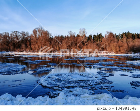 Frozen River with Floating Ice Floes in Winter. Wide view of partially frozen river surface covered with ice chunks under clear sky with forested shore in background. Frozen River with Floating Ice Floes in Winter. Wide view of partially frozen river surface covered with ice chunks under clear sky with forested shore in background. 134991348