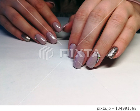 Purple Glitter Accent Manicure. Hands displaying manicured nails in lavender shades with silver glitter accents, rhinestones and glossy top coat on white background. Purple Glitter Accent Manicure. Hands displaying manicured nails in lavender shades with silver glitter accents, rhinestones and glossy top coat on white background. 134991368