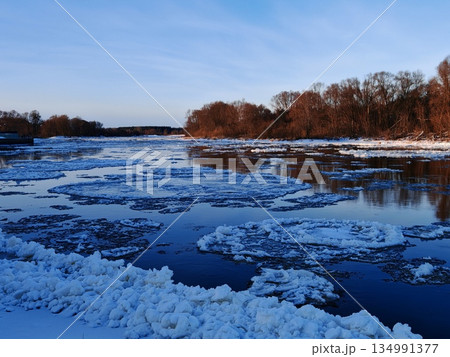 Winter River Landscape with Floating Ice Floes. Wide view of river partially covered with drifting ice chunks under clear blue sky with distant tree line. Winter River Landscape with Floating Ice Floes. Wide view of river partially covered with drifting ice chunks under clear blue sky with distant tree line. 134991377