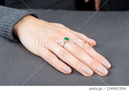 Beautiful female hand with gold ring with diamonds and emerald on gray background Beautiful female hand with gold ring with diamonds and emerald on gray background 134991584