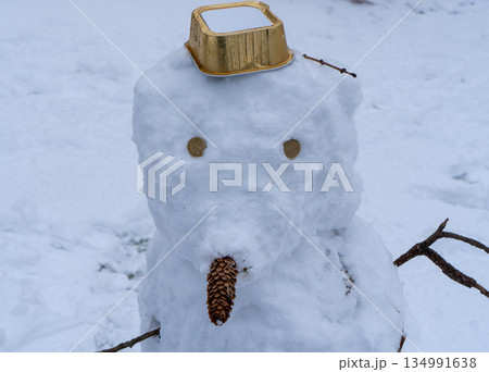 Macro view of a snowman's face made from white snow. Features include eyes made of small objects, a nose made from a pine cone, and a gold-colored plastic container as a hat. Blurred snow background. 134991638