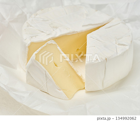 A round head of soft Camembert cheese with white mold, one triangular slice cut off, the cheese lies on crumpled white wrapping paper 134992062