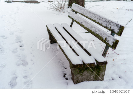 An empty wooden bench with a backrest sits in a winter park, covered with a thick layer of fresh snow. Footprints are visible on the snow-covered ground nearby. Natural daylight, overcast winter day. An empty wooden bench with a backrest sits in a winter park, covered with a thick layer of fresh snow. Footprints are visible on the snow-covered ground nearby. Natural daylight, overcast winter day. 134992107