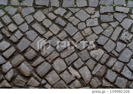 High-angle view of a grey stone pavement made of irregular cobblestones. Wet masonry texture with some green moss in cracks 134992108