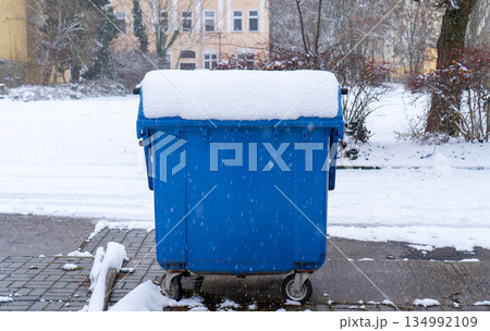 large blue industrial trash bin with wheels stands outdoors on a snowy day. A thick, smooth layer of fresh white snow covers the lid. City street and buildings in the background. large blue industrial trash bin with wheels stands outdoors on a snowy day. A thick, smooth layer of fresh white snow covers the lid. City street and buildings in the background. 134992109