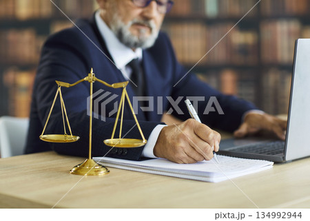 Attorney writing notes at desk with laptop and justice scales in law library Attorney writing notes at desk with laptop and justice scales in law library 134992944
