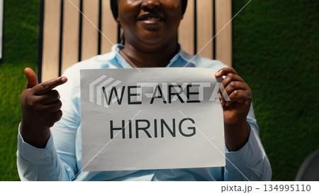 African american female HR manager showing recruiting signs on papers to create a talent acquisition, announcing job vacancy and inviting skilled people to join the company workforce. African american female HR manager showing recruiting signs on papers to create a talent acquisition, announcing job vacancy and inviting skilled people to join the company workforce. 134995110