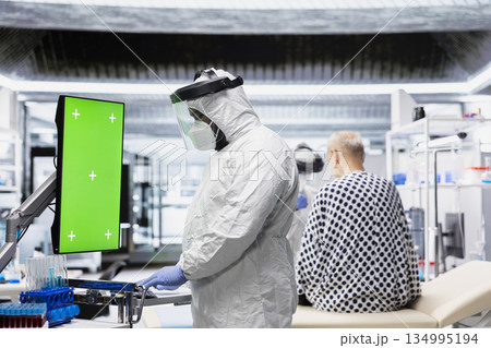 Vertical chroma key monitor next to scientist in protective gear examining virus symptoms of patient in prevention center. Safety data and breakthrough treatment results for bioscience. 134995194