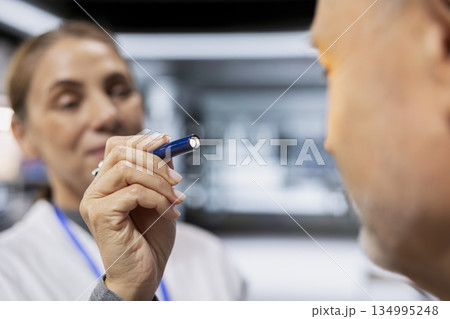 Close up of female expert keeping senior man under observation in clinic laboratory, checking potential side effects during new drug testing with light pen. Dosage safety for medical progress. 134995248