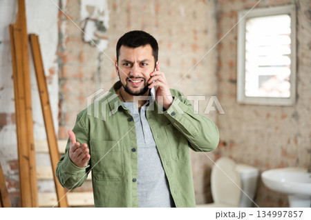 Dissatisfied bearded man talking on phone in house during renovation Dissatisfied bearded man talking on phone in house during renovation 134997857