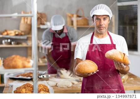 Caucasian male cook-baker worker with loafs of bread, shows many products Caucasian male cook-baker worker with loafs of bread, shows many products 134997871
