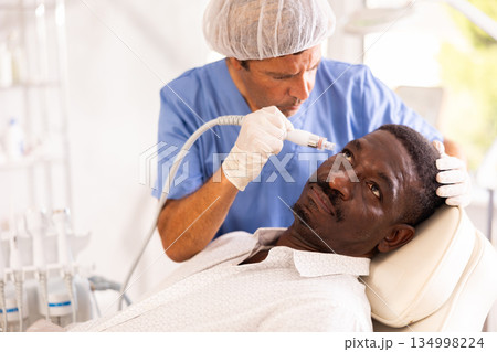 Male cosmetologist performs skin cleansing procedure for African man client with beauty machine Male cosmetologist performs skin cleansing procedure for African man client with beauty machine 134998224
