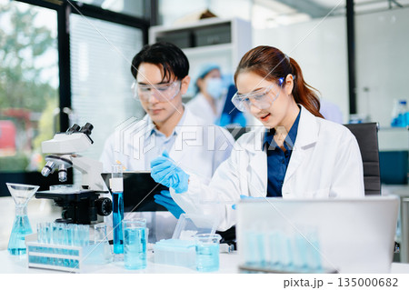 Scientists collaborating in a modern lab with microscopes and blue liquids, representing biotechnology, chemistry, and medical research innovation. 135000682