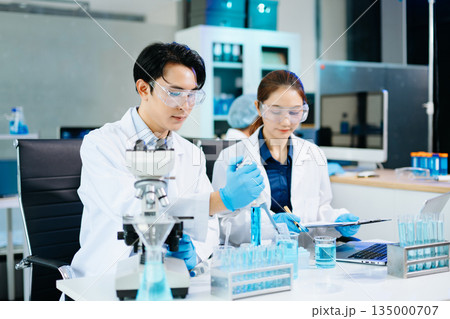 Scientists collaborating in a modern lab with microscopes and blue liquids, representing biotechnology, chemistry, and medical research innovation. 135000707