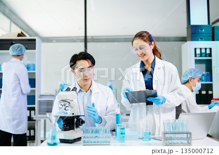 Scientists collaborating in a modern lab with microscopes and blue liquids, representing biotechnology, chemistry, and medical research innovation. 135000712