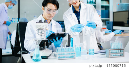 Scientists collaborating in a bright modern lab with blue liquids and microscopes, symbolizing teamwork, biotechnology, chemistry, and medical innovation. 135000716
