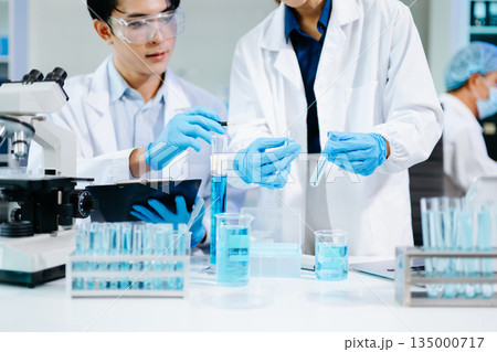 Closeup of Asian scientists experimenting with blue liquids and microscopes in a modern lab, representing biotechnology, chemistry, and medical innovation. 135000717