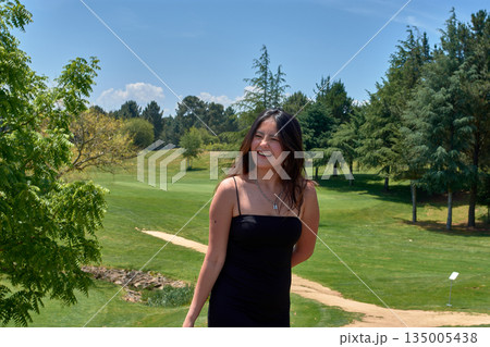 A young woman in a black dress smiles happily as she poses on a sunny golf course surrounded by pine trees and green grass under a blue sky A young woman in a black dress smiles happily as she poses on a sunny golf course surrounded by pine trees and green grass under a blue sky 135005438