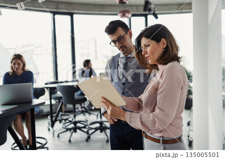 Two collegues talking about project document, standing in modern office. 135005501