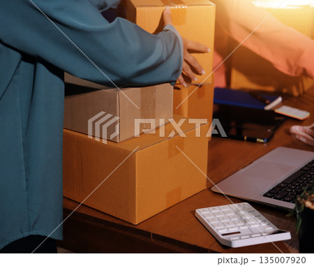 Startup SME small business entrepreneur of freelance Asian woman wearing apron using laptop and box to receive and review orders online to prepare to pack sell to customers, online sme business ideas. Startup SME small business entrepreneur of freelance Asian woman wearing apron using laptop and box to receive and review orders online to prepare to pack sell to customers, online sme business ideas. 135007920