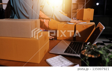 Startup SME small business entrepreneur of freelance Asian woman wearing apron using laptop and box to receive and review orders online to prepare to pack sell to customers, online sme business ideas. 135007921