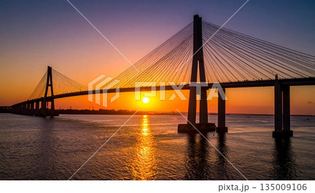 A vibrant sunset and sunrise reflect over the city river and calm lake water near the bridge and harbor pier as clouds fill the summer sky at dusk A vibrant sunset and sunrise reflect over the city river and calm lake water near the bridge and harbor pier as clouds fill the summer sky at dusk 135009106
