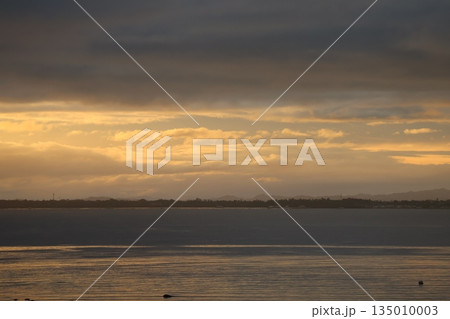 雲の多い朝焼けのオレンジ色の空と対岸の海岸と霞む山と穏やかな海面の海の風景 雲の多い朝焼けのオレンジ色の空と対岸の海岸と霞む山と穏やかな海面の海の風景 135010003