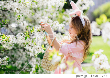 Easter egg hunt. Girl child Wearing Bunny Ears Running To Pick Up Egg In Garden. Easter tradition. Baby with basket full of colorful eggs. Easter egg hunt. Girl child Wearing Bunny Ears Running To Pick Up Egg In Garden. Easter tradition. Baby with basket full of colorful eggs. 135011747