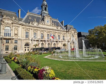 The Hotel de Ville in Tours, France. Grand Neoclassical building with ornate facade and classic clock tower, decorated with beautiful tiered fountain and lush green garden with vibrant flower beds 135012605