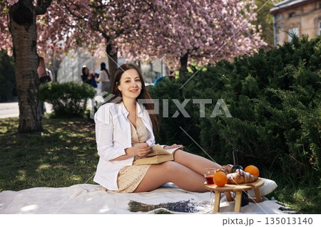 spring scene, picnic setup, seasonal fruit. Sitting on picnic blanket under blooming cherry trees, young woman enjoys book and holds flower, with snacks and fruit nearby. spring scene, picnic setup, seasonal fruit. Sitting on picnic blanket under blooming cherry trees, young woman enjoys book and holds flower, with snacks and fruit nearby. 135013140