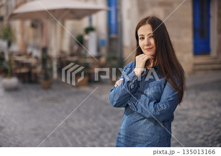 Denim fashion, City background, Denim dress. Young woman stands smiling in denim dress on cobblestone street, enjoying casual urban day. 135013166