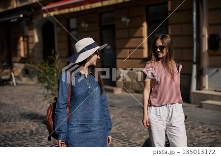 walking down street, happy travelers, street travel. Carrying suitcases, two casually dressed young women walk down cobblestone street enjoying sunny urban atmosphere. walking down street, happy travelers, street travel. Carrying suitcases, two casually dressed young women walk down cobblestone street enjoying sunny urban atmosphere. 135013172