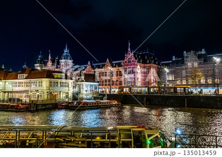 Amsterdam Railway station canal water house building. Netherlands night. Evening street architecture. Water boats Amsterdam Railway station canal water house building. Netherlands night. Evening street architecture. Water boats 135013459